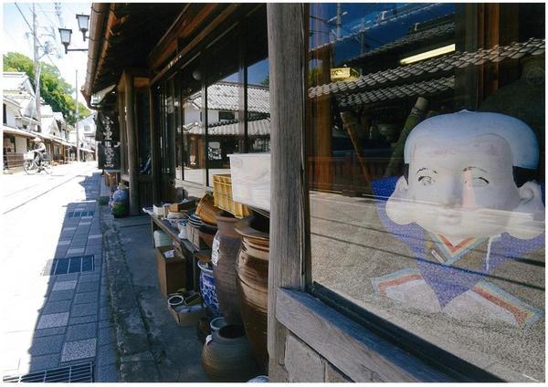 旧街道の家の軒下に様々な形の壺やガラスケース内に置かれた置物の写真