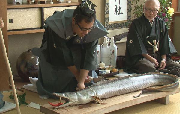衣装を着た男性が、大きな鱧を切りつけている写真