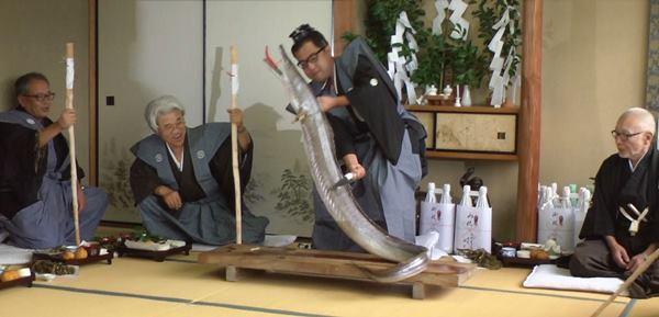 衣装を着た男性が、大きな鱧を持ち上げながら切りつけているのを衣装を着た男性2名が棒をもっている写真