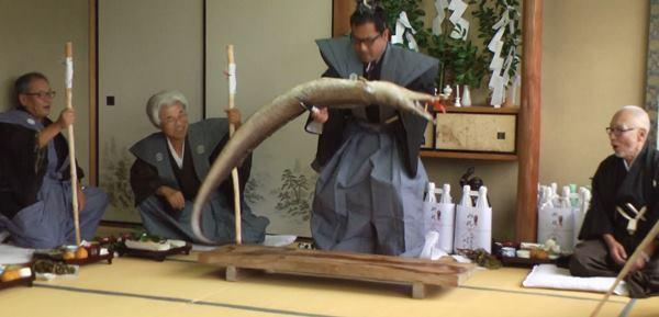 衣装を着た男性が、大きな鱧を腹を上に逆さに持ち上げ、その横で、男性2名が見ている写真