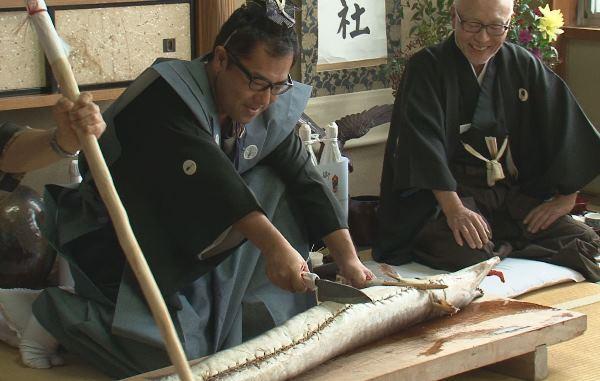 衣装を着た男性が、お腹を縫われた大きな鱧に、包丁をあてている写真