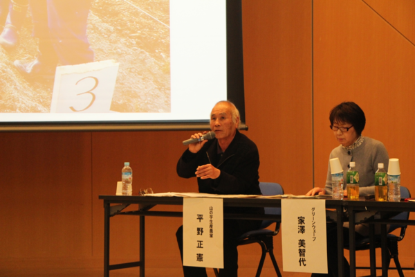 スクリーンに映像が映っており、その横で椅子に座りながらマイクを持った平野 正憲さんが発表している横で資料を見ている家澤 美智代さんの写真
