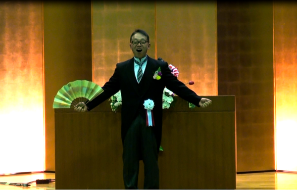 丹後校長先生が舞台の前で扇子を持ちながら手を広げ、口を大きく開けて「ワッハッハ！」と言っている写真