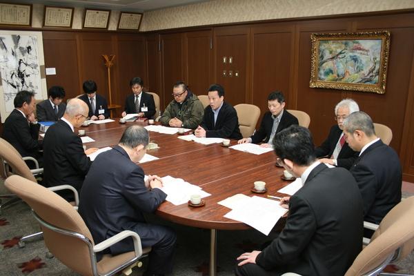 楕円形のテーブルの周りに11名のスーツを来た男性と市長が座って資料を見ながら話をしている写真