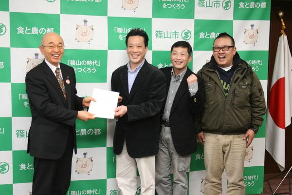 篠山市と書かれた壁の前で右はじにメガネの男性、その左隣にガッツポーズをした男性、その右隣に黒いジャケットの男性と市長が白い紙を二人で持っている写真