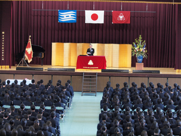 ステージ上の壇上で男性が卒業生と在校生に向かって話しをしている写真