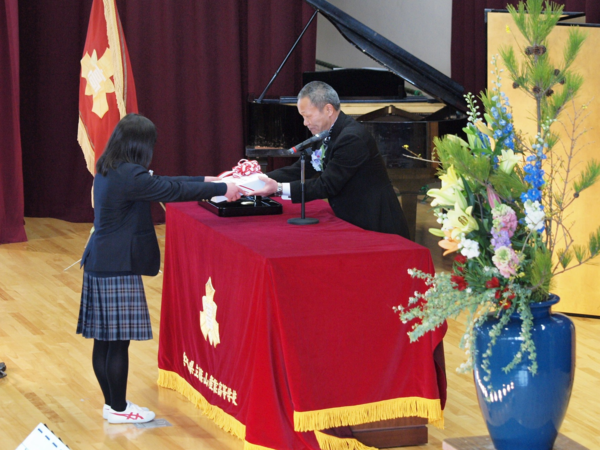 ステージ上で男性が卒業生代表の女子生徒へ卒業記念品を手渡している写真