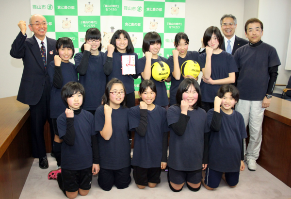 ガッツポーズをしている市長と、男性2名、がライズ篠山ドッジボールクラブの女子生徒達をはさみ前列の5人が膝たちをして後列の二人がボールをもち一人が奨励金を手に持ちみんなで子供が右手を曲げ前にだしポーズをしている写真