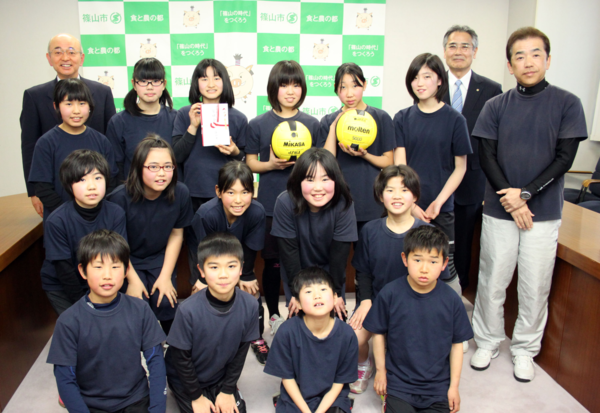 1列目に男の子4人が座り2列目に女の子5人が膝に手をおき中腰、3列目に女の子6人がたちボールを2個もち奨励金をもち周りに市長と男性2人が並びみんなでの集合写真
