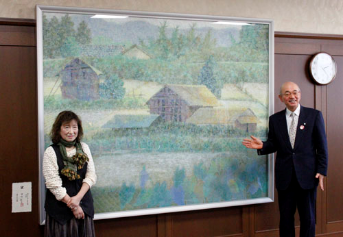 応接室に飾られた、大きな農村風景の絵画の前に立つ市長と女性の写真