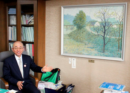 市長室に飾られた、農村風景の絵画のまえに座る市長の写真