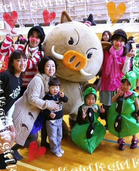 まるいの両隣に鼻にボンボンをつけた人がポーズをとりその周りにピースをした緑の衣装の子供や子供と大人が集まって一緒に記念写真