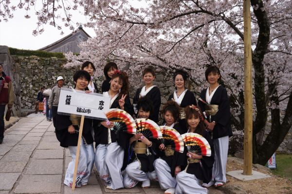 よさこい踊りに参加した白色のズボンを履いて黒色の羽織を着た女性のチームが桜の木の下で扇子を手にポーズをとっている集合写真
