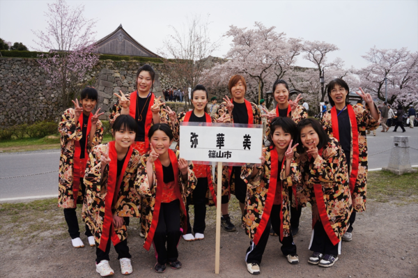 黒地に金色をベースにした花柄の法被を着た「弥華美」チームが会場の桜並木の前でピースサインをしている集合写真