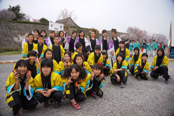 黄色の法被を着た子どもと右袖が白地に紫色の柄の法被を着た大人が混在したチーム「北翔」が桜並木の前の集合写真