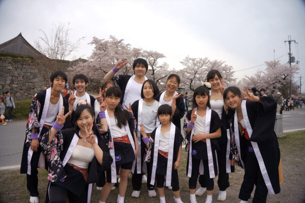 右袖のみ着物の花柄が入っている黒色の法被を着た男女混合のチーム「輝楽」メンバーが桜並木の前でピースサインをしている集合写真