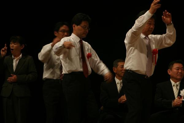 篠山市の職員が上着を脱いで、デカンショ節を踊っており、後方の男性は手拍子をしている様子の写真