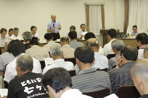 前列の一人がマイクを持ち参加者に説明をしているのをたくさんの人が椅子に座り聞いている写真