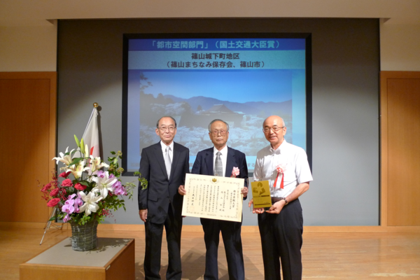 スクリーンの前に男性3人が立って中央の男性が賞状を市長が盾をもって記念写真
