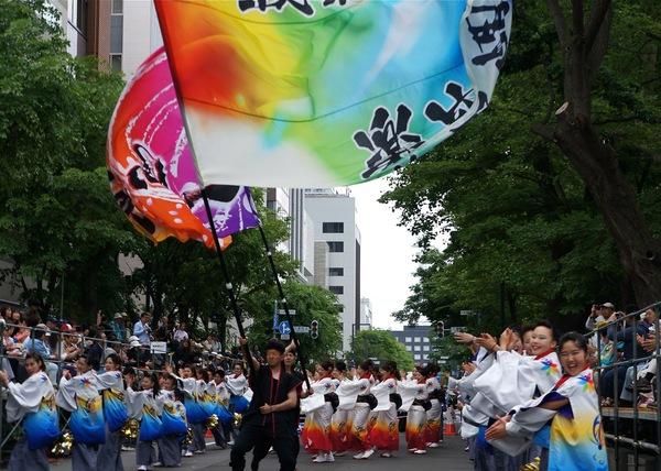 緑の街路樹が並ぶ道路で大漁旗のような大きな旗を振っている男性2名とその周りをはっぴや浴衣を着た丹波篠山楽空間一期一会の皆さんが踊っている写真