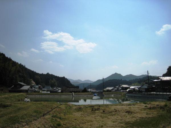 山々の麓に住宅が並び、中央にため池があり、手前に田んぼが写っている写真