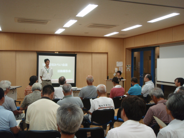 篠山環境みらい会議の活動報告会会場でスクリーンの前で男性が立って話しをし、在籍する委員が椅子に座って聞いている後ろ姿の写真
