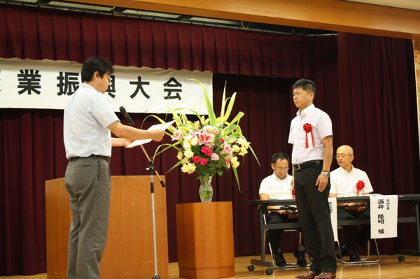 舞台に上がって、胸に赤い花の胸章リボンを付けた酒井 一行さんが表彰をされている様子の写真