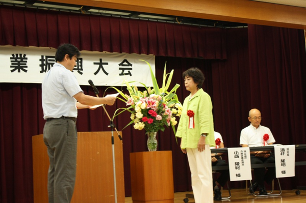 舞台に上がって、胸に赤い花の胸章リボンを付けた波多野 ふみ子さんが表彰をされている様子の写真