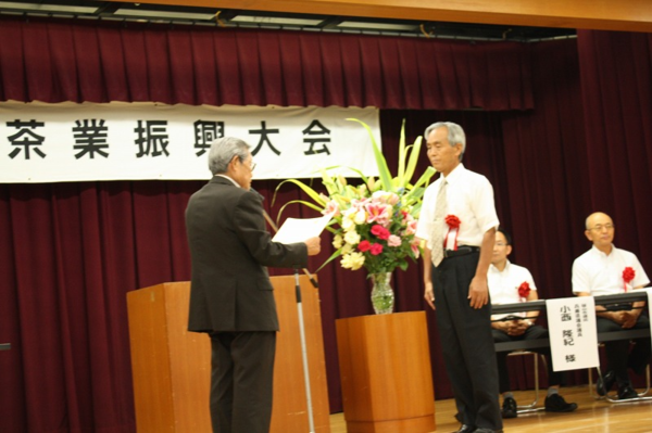 舞台に上がって、胸に赤い花の胸章リボンを付けた柳澤さんが表彰をされている様子の写真