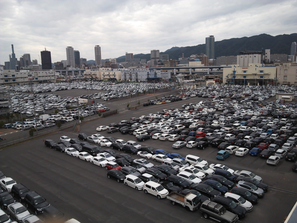 奥にはビル街があり、手前には100台以上の車が停めている駐車場の空撮写真