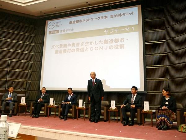 スクリーンのある舞台上には6名の方が席についており、市長が席から立ちあがって一礼している様子の写真