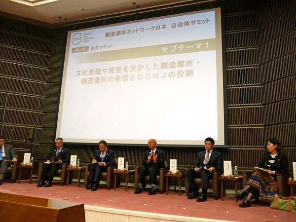 スクリーンのある舞台上で6名が席についており、市長がマイクを握って話をしている様子の写真