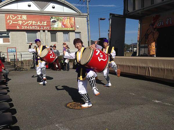 パン工房ワーキングたんば＆スマイルカフェふわりの看板の前の道路で3人の人がはっぴを着て、左足をあげながら赤い太鼓を叩いている写真