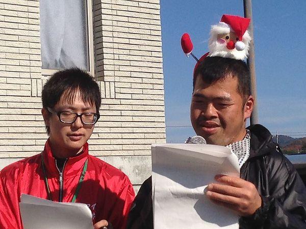 男性2人が資料を手に持ち、1人はサンタの人形の帽子を被ってマイクを持ってお話ししている写真