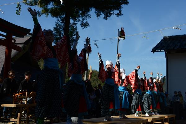 うす暗い神社の鳥居の前で頭に白い被り物をし、赤と水玉模様の衣装を着た人々が両手を上にあげている写真
