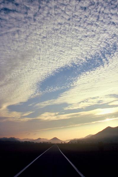 空が白い雲で覆われ、遠くの山並みには光が差している風景を道路から真正面に見た写真
