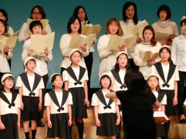 合唱団の方と子供達が女性の指揮者に合わせて合唱している写真