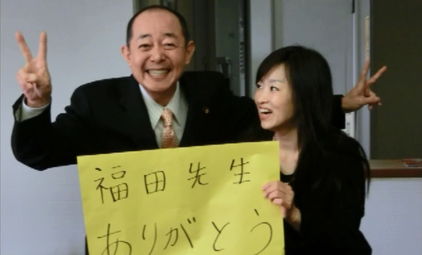 女性が「福田先生ありがとう」のメッセージカードを持ってその隣で男性が笑顔でピースしている写真
