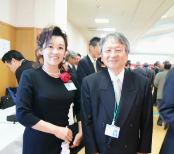 眼鏡をかけた男性と正装した女性が2人並んで笑顔で写っている写真