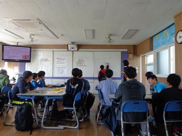 授業で生徒がホワイトボードに書いている答えを見ている先生と生徒たちの写真