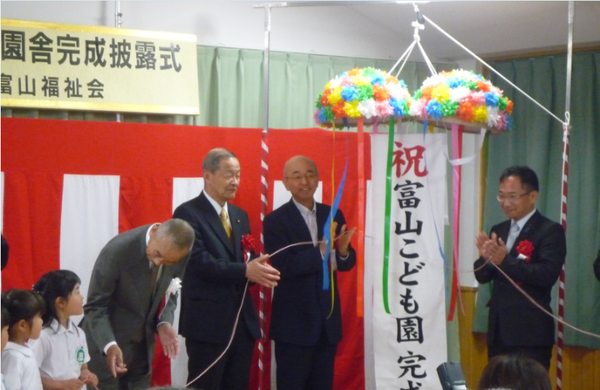 富山こども園の完成披露式でくす玉が割られ拍手をしている市長や三重野理事長らと園児らが写っている写真