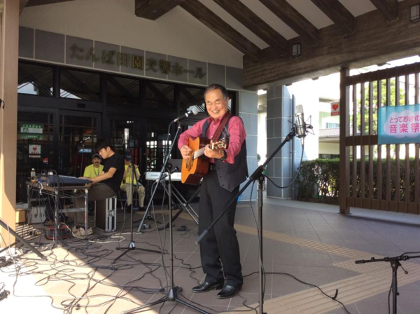 たんば田園交響ホールの外で男性がスタンドマイクの前に立ちギターを演奏している写真