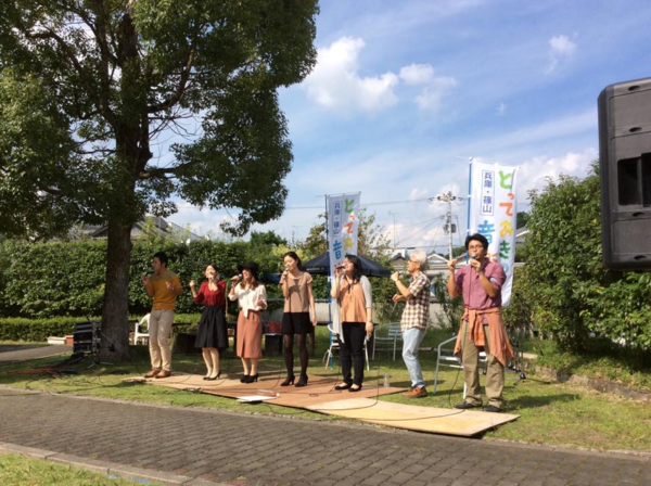 芝生の木陰の所で男女7名がマイクを手に持ち歌っている写真