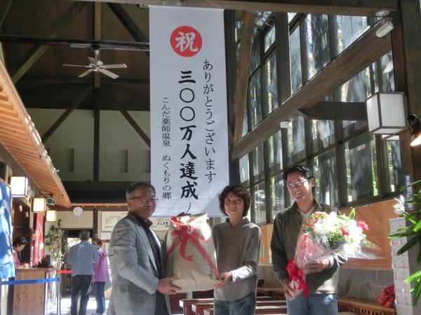 300万人達成の垂れ幕の下で、花を持つ男性と、男性と女性が一緒に、リボン柄が印刷された米袋を持っている写真