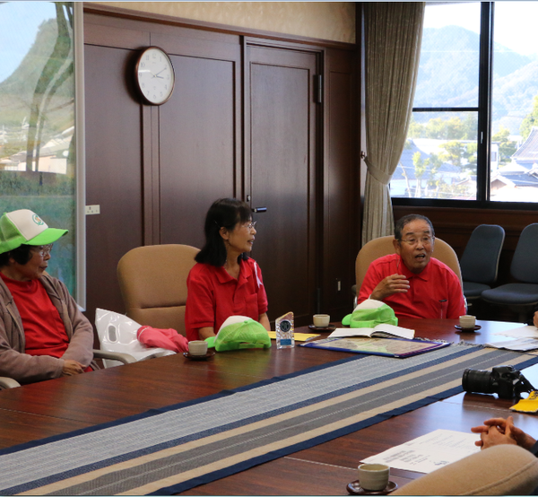 長机に座っている卓球バレーの男性メンバーが話をしており、それを他のメンバー聞いている写真