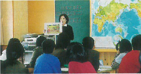 黒い服を着た女性が教壇に立ち右手に拡大した写真を手に持ち話しをしているのを聞いている生徒の後ろ姿の写真