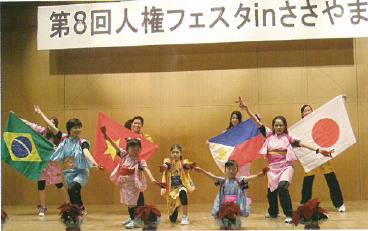 人権フェスタinささやまのステージ上で着物を着てタスキで袖をくくりズボンを履いた格好で前方で右手を挙げ左手を下に向けて両手を大きく開いた踊りの決めポーズをとり、後方では一人ずつ4か国の国旗を手に持った女性の写真