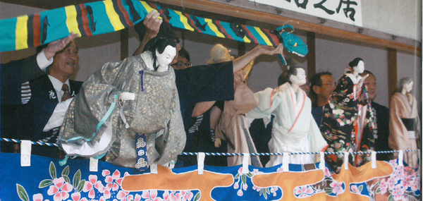 舞台の上で着物を着た人形を後ろで男性が操って人形狂言をしている様子の写真