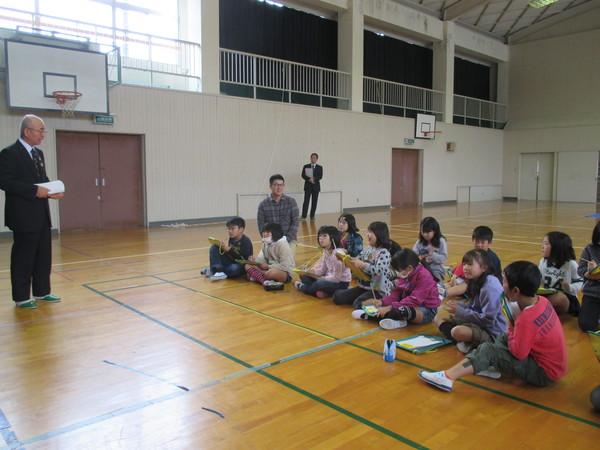 体育館で市長が床に座っている3年生に話をし、児童たちはメモを取っている様子の写真