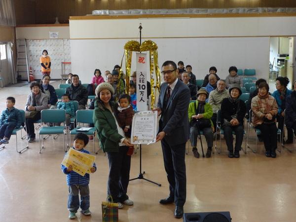 来館者数が10万人に到達でゴールドのくす玉と垣内館長からお祝いされる村上さん親子とお客様の写真